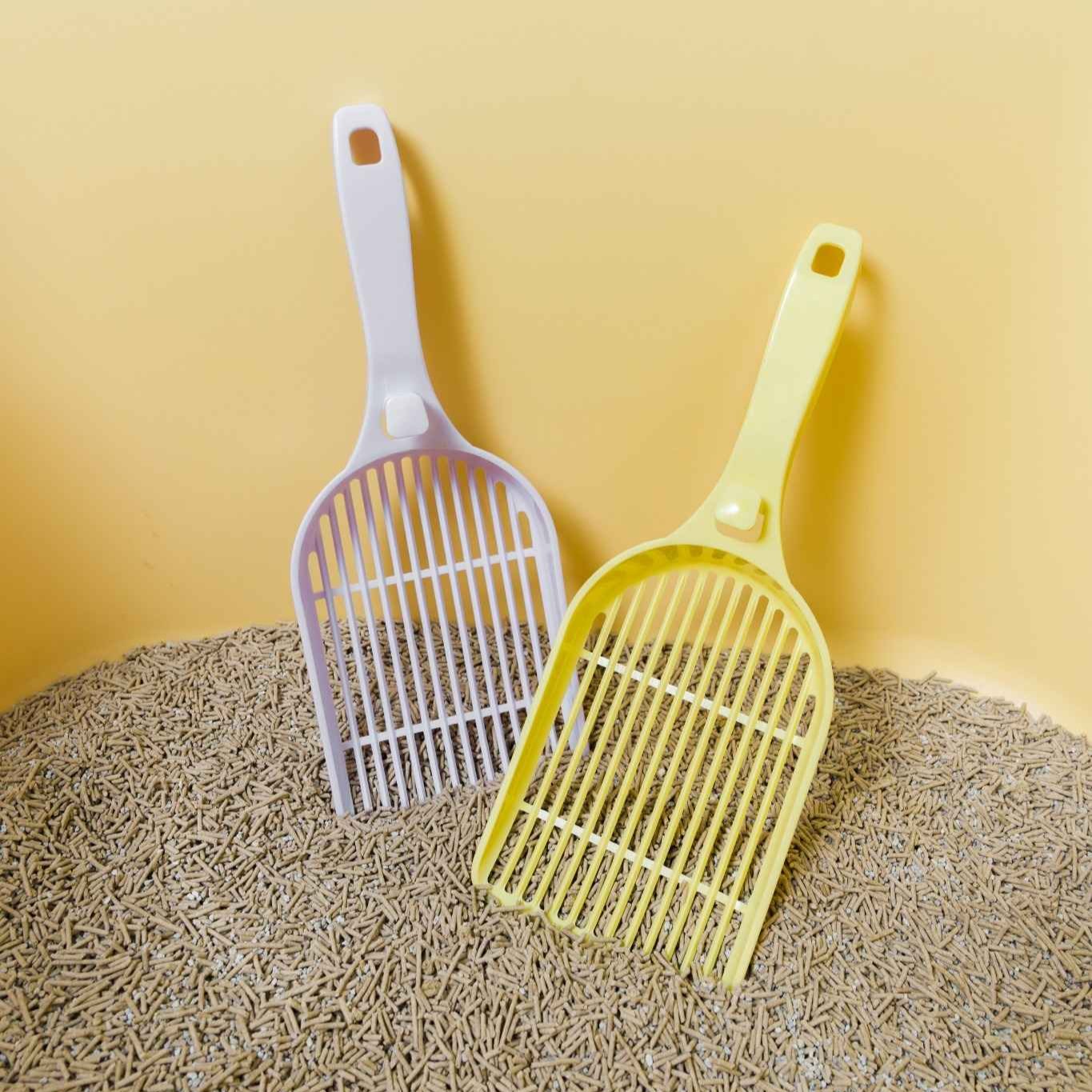 Two pet litter scoops, one white and one yellow, on a bed of cat litter against a yellow background.